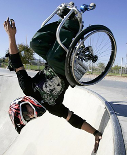A wheelchair rider goes fully inverted at a skate park – chair overhead, arms extended, helmet on