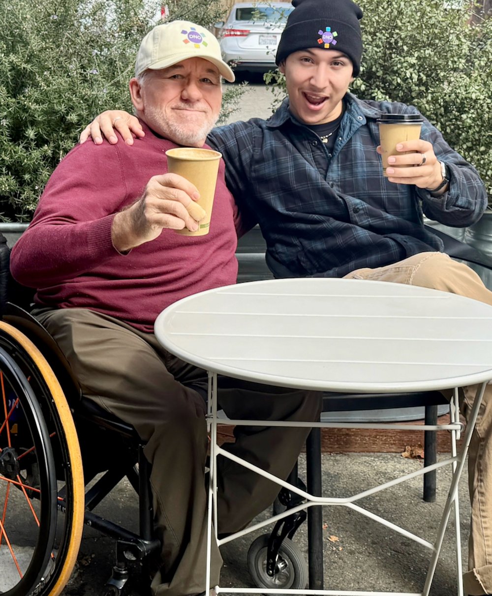 Erick Mikiten, in his wheelchair and a DNO cap, toasting coffee cups outdoors with his son's friend Aiden, who wears a DNO beanie