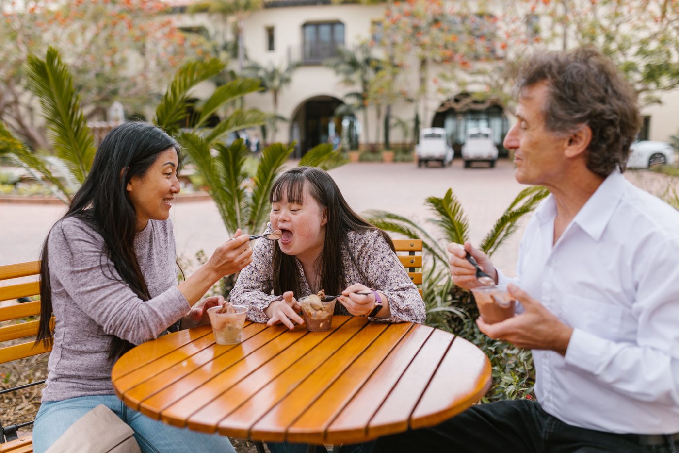 A young woman with Down syndrome opens wide for a spoonful of dessert held by the woman beside her, while a man on her other side smiles, at an outdoor cafe table.