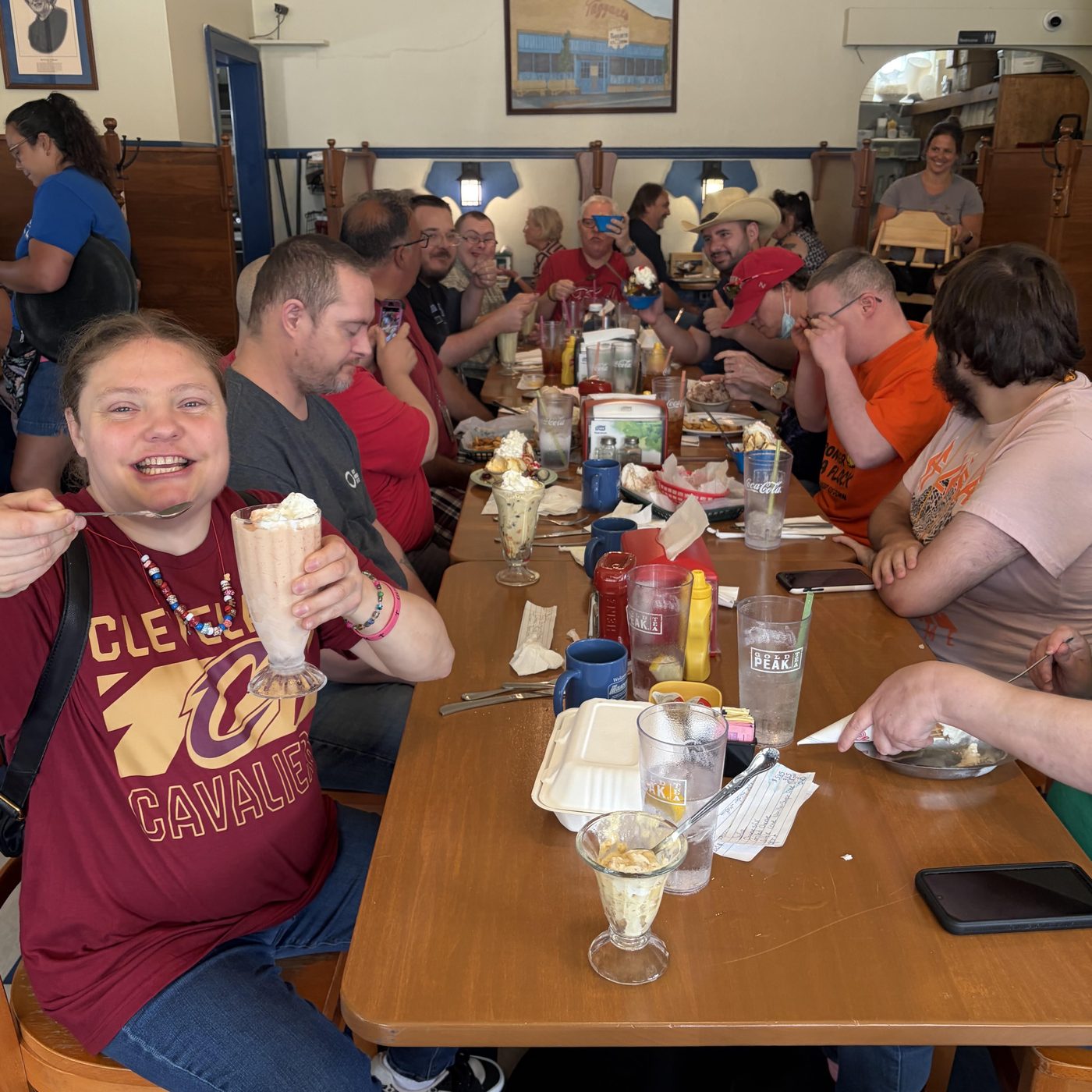 A long table of diners in a classic American diner, laughing and sharing milkshakes and sundaes; a woman in a Cleveland Cavaliers shirt in the foreground holds up a milkshake and smiles at the camera.
