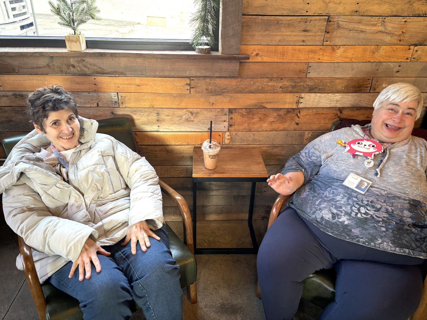 Two women sit and smile in armchairs inside a cafe with wooden walls; a drink with a straw is on a small table between them.