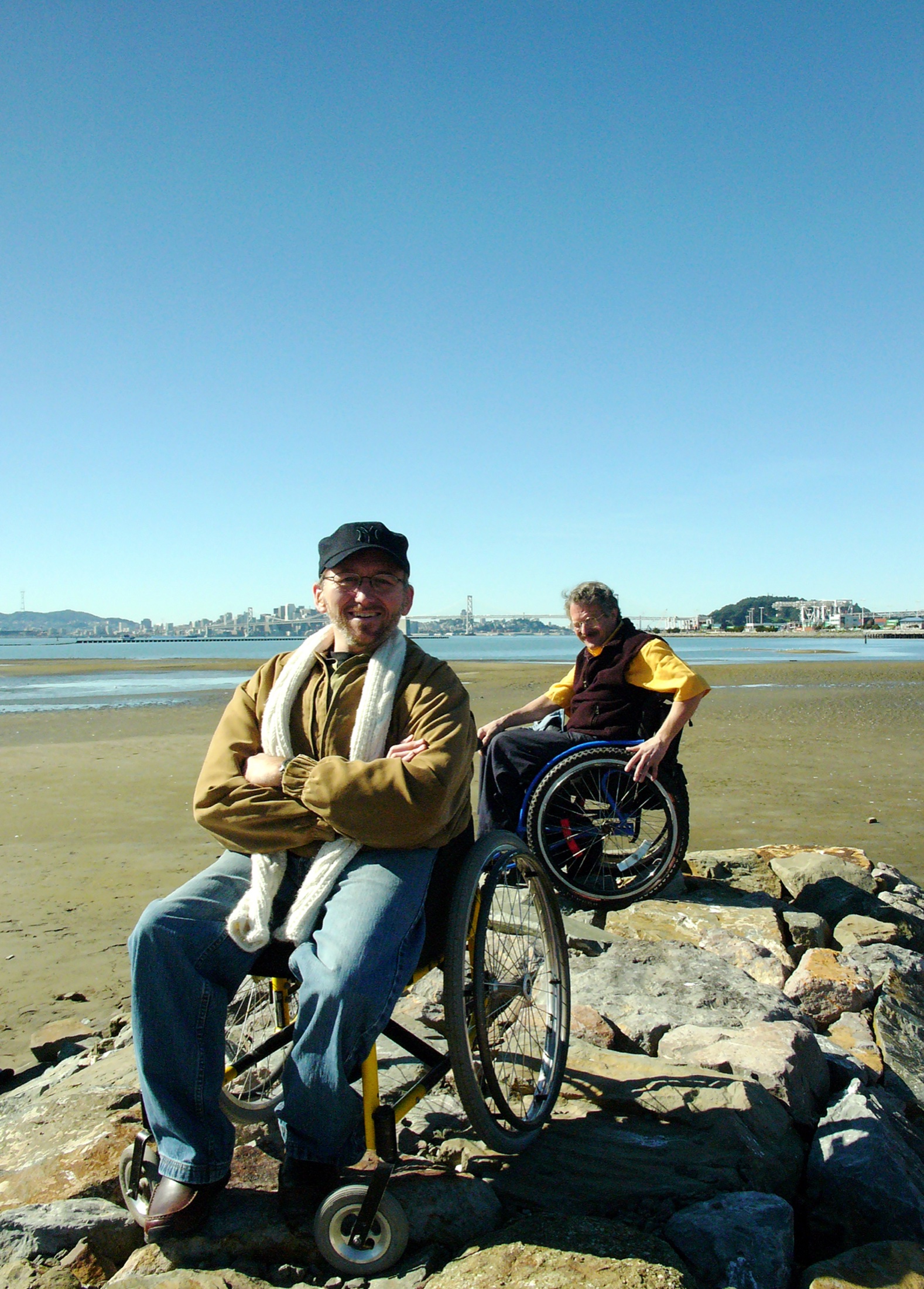 Erick and a friend, both in wheelchairs, on rocks at Alameda Beach with the Bay Bridge behind them