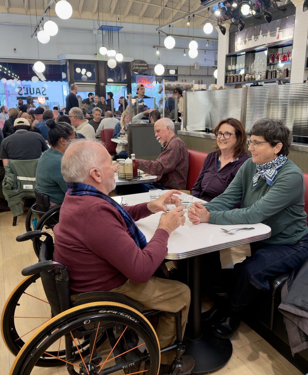 Erick Mikiten, an architect and wheelchair user, holding hands across the table with his friends Elisa and Libby at a busy, crowded restaurant